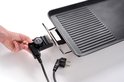 Een hand draait aan de temperatuurregelaar van een elektrische grillplaat. Een afneembare stekker ligt ernaast. Het gladde grilloppervlak is zichtbaar.