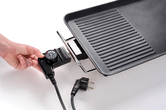 Een hand draait aan de temperatuurregelaar van een elektrische grillplaat. Een afneembare stekker ligt ernaast. Het gladde grilloppervlak is zichtbaar.