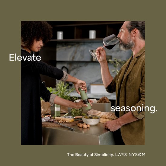 Vrouw en man aan een keukenblad. Vrouw gebruikt een groene molen. Man drinkt uit een glas rode wijn. Tekst: Elevate seasoning.
