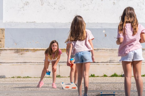 Meisjes spelen cornhole, met blauwe en oranje beanbags. Eén meisje kijkt naar voren, de ander naar het spel.