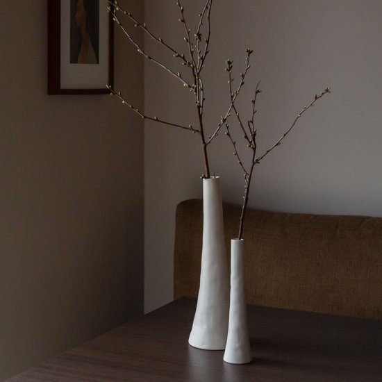Twee slanke, crèmekleurige keramieken vazen met takken op een donkere tafel.