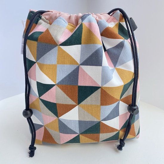 Grijze projecttas met geometrisch patroon van roze, oker, terracotta, groen en grijze driehoeken. Zwarte trekkoorden met stoppers.