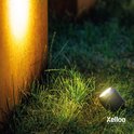 Een zwarte Xelloo grondspot verlicht gras en een roestige metalen paal bij nacht.