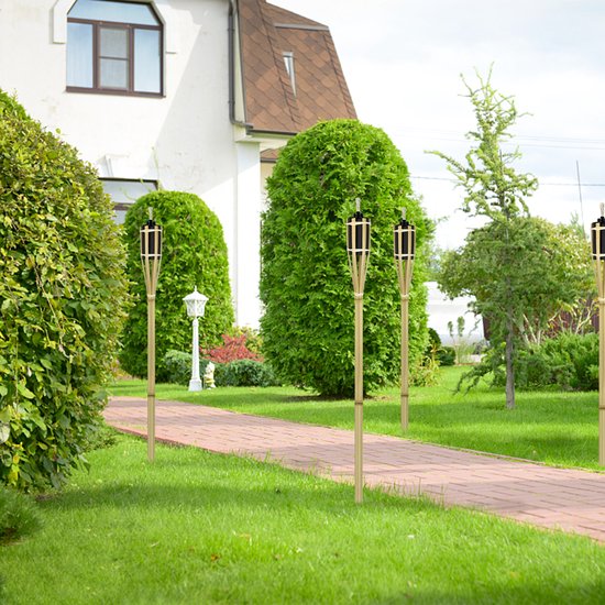 Pad in een tuin met vier bamboe tuinfakkels met vlammen, langs een stenen pad en groen struikgewas.