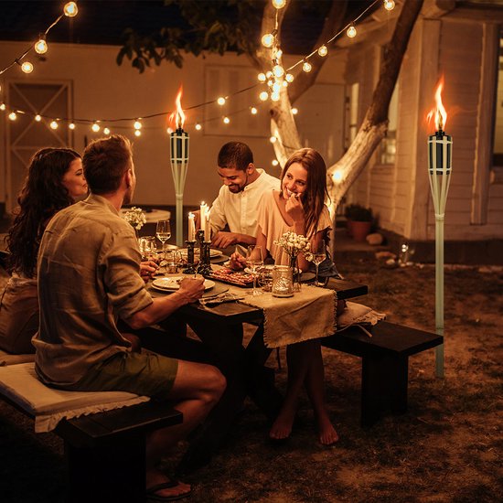 Dinerende vrienden aan een picknicktafel 's avonds, met twee brandende bamboe tuinfakkels op de achtergrond.