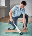 Man knielt op een groene ondergrond en gebruikt een laminaatsnijder om een houten plank door te snijden.