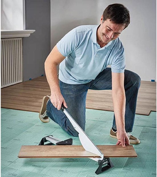 Man knielt op een groene ondergrond en gebruikt een laminaatsnijder om een houten plank door te snijden.