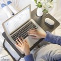 Vrouw op bed met laptop op een donker houtnerf laptoptafel, met een witte muis en vaas met bloemen.