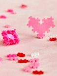 Een roze Plus-Plus hart en een gebalde Plus-Plus vorm in roze. Losse rode, roze en witte steentjes liggen op de achtergrond.