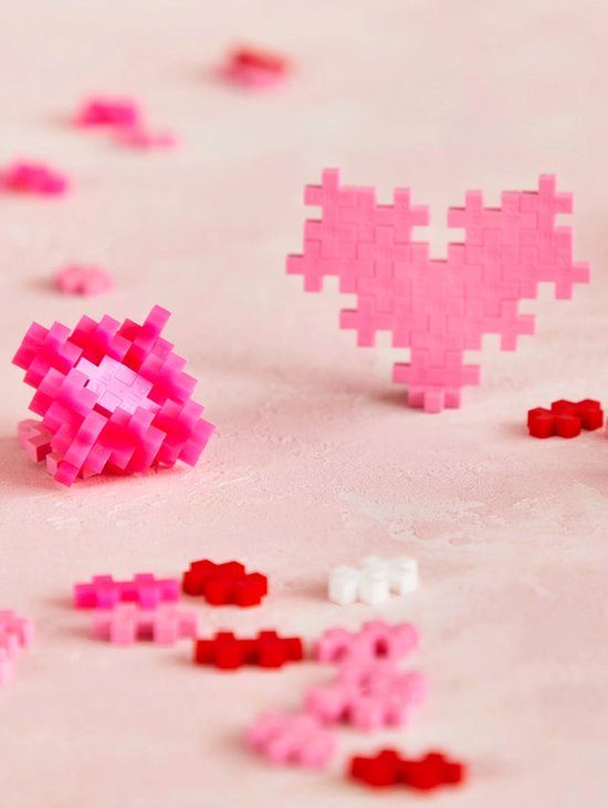 Een roze Plus-Plus hart en een gebalde Plus-Plus vorm in roze. Losse rode, roze en witte steentjes liggen op de achtergrond.