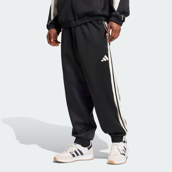 Man in zwarte trainingsbroek met witte strepen en Adidas logo, gedragen met witte sneakers.