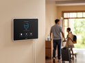 Eufy smart display op een muur met "Away" instelling en een gezin dat vertrekt. Wit display met zwarte rand en rode accenten.