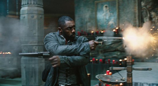 Idris Elba in donkergrijze jas, vuurt twee pistolen af te midden van kaarsen en rook.