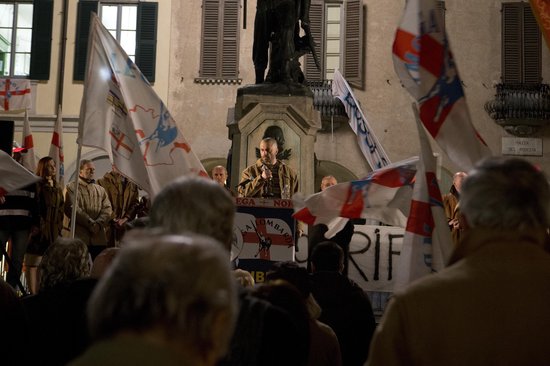 Een man spreekt tot een menigte voor een standbeeld, omringd door Italiaanse vlaggen.