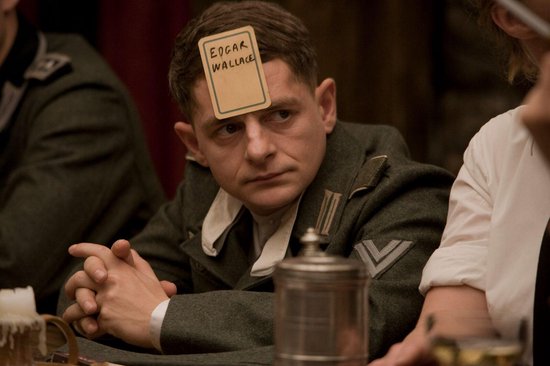 Een man met een kaart met 'EDGAR WALLACE' op zijn voorhoofd, gekleed in een uniform, zit aan tafel met gevouwen handen.