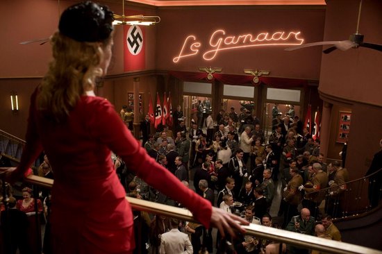 Een vrouw in een rode jurk kijkt uit over een drukke menigte in een theaterzaal, versierd met nazi-vlaggen en een neonbord.