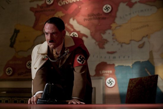 Een man met snor, gekleed in een bruin uniform met een cape en een swastikaband om zijn arm, leunt op een bureau met een kaart.