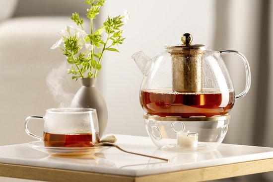 Glazen theepot met gouden filter, verwarmd door een kaarsje op een glazen onderzetter. Een kopje thee staat ernaast.