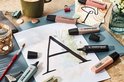 Meerdere STABILO BOSS ORIGINAL stiften, waaronder zwart, pastelgroen en roze, rond een tekening van de letter 'A'.
