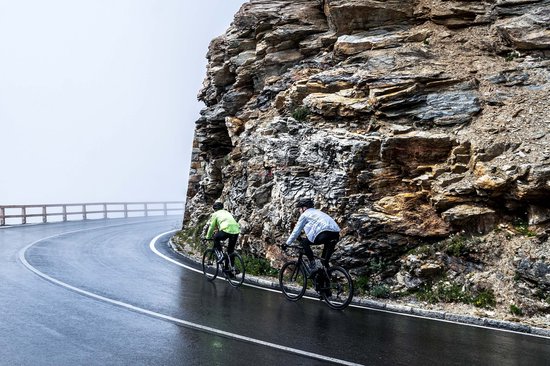 Twee fietsers op een natte, bochtige bergweg. Eén in een witte jas, de ander in een neongele jas.