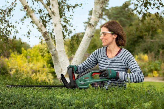 Vrouw met groene en zwarte heggenschaar met rode accenten, snoeit een heg.