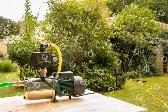 Zilverkleurige hydrofoorpomp met groene motorbehuizing en zwarte besturingseenheid, met gele en groene slang, in een tuin op een houten tafel.
