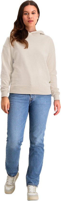 Vrouw met bruin haar, gekleed in een crème hoodie en spijkerbroek, met het logo op de heup.