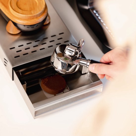 Een hand houdt een zilverkleurige portafilter met houten handvat boven een uitklaplade van geborsteld staal.