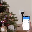 Een slimme stekker aangesloten op een stopcontact naast een kerstboom. Een telefoon toont de tijdschema-instellingen.