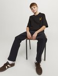 Jongen zit op een stoel, draagt een zwarte t-shirt met gele tekstprint en donkergrijze cargo broek.