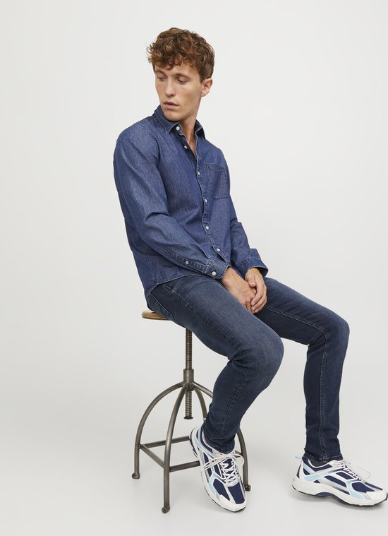 Zittende man in blauwe skinny jeans en spierwitte denim blouse, kijkend naar links.