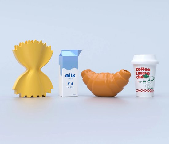 Oranje keramieken vaas in de vorm van een croissant, naast een gele farfalle-vormige vaas, melkpak en koffiebeker.