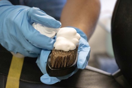 Gants bleus pulvérisant une mousse blanche sur une brosse foncée à poils bruns.