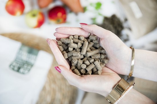 Twee handen met daarin een hoopje koudgeperste hondenbrokken, op de achtergrond appels en wortels.