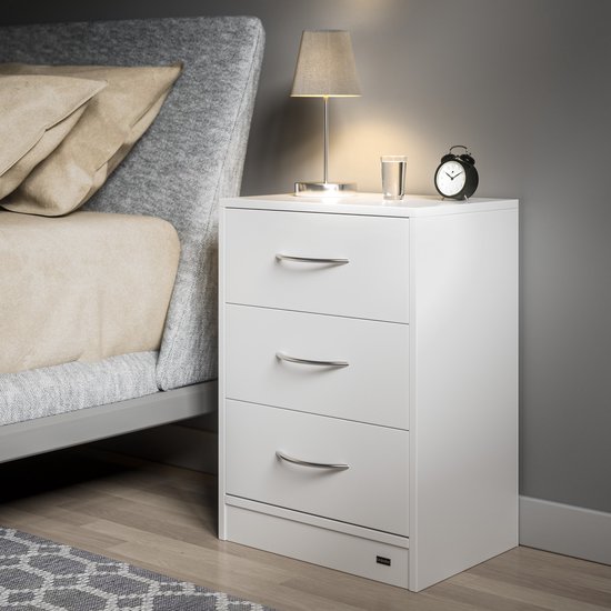Wit nachtkastje met drie lades naast een grijs bed. Een tafellamp en wekker staan op het nachtkastje.