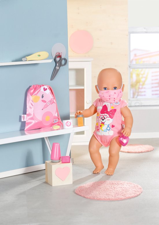 Een baby Born pop met masker in een roze outfit, houdt een thermometer vast.