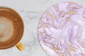 Sous-verre rond à motif marbre lilas et or, à côté d'une tasse de café.