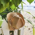 Bruine strooien cowboyhoed met schelpenband, hangend aan een wit hekje bij de zee.
