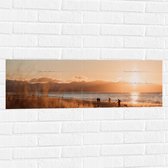 WallClassics - Muursticker - Zon Verdwijnend achter Bergen bij Strand - 90x30 cm Foto op Muursticker