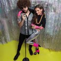 Man met afro, zonnebril en disco-shirt zingt in microfoon. Vrouw met felgekleurde accessoires, glittertop en metallic broek.