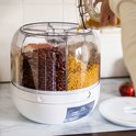 Witte voorraaddispenser met vier vakken gevuld met bonen, pasta en granen. Een hand vult pasta bij.