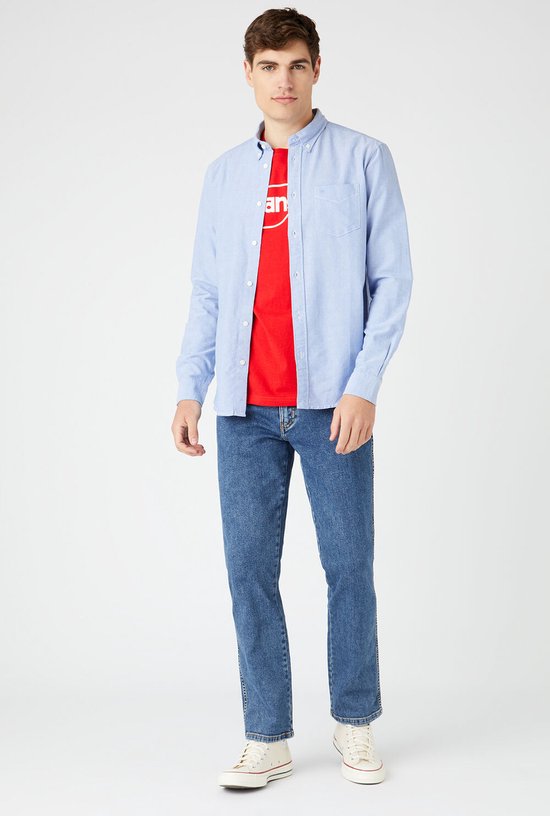 Man, staand, draagt een blauwe spijkerbroek met een rode Wrangler t-shirt en blauw overhemd.