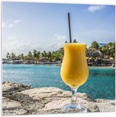 WallClassics - PVC Schuimplaat- Gele Cocktail op Rots aan het Water van Vakantieplek - 100x100 cm Foto op PVC Schuimplaat