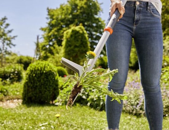 GARDENA onkruidsteker houdt paardenbloem met wortel omhoog, vrouw in jeans op achtergrond.