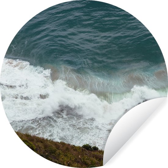Zelfklevende ronde muursticker met een luchtfoto van brekende golven aan een kustlijn, met een loslatende hoek.
