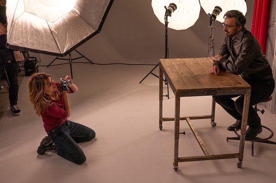 Vrouw met camera fotografeert man aan tafel in studio met verlichting.