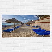 WallClassics - Muursticker - Blauwe Ligbedden op Strand met Rieten Parasols - 100x50 cm Foto op Muursticker