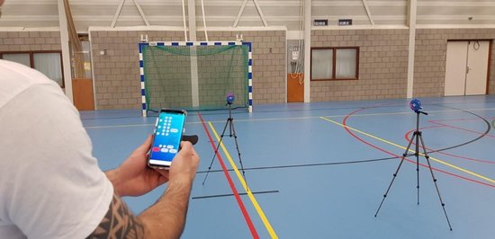 Man met getatoeëerde arm houdt smartphone vast en bedient twee blauwe reaction lights op statieven in sporthal.