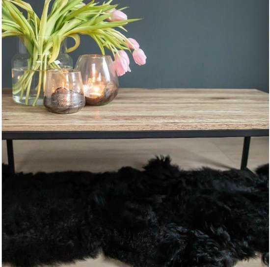 Salontafel met licht houten blad en zwart stalen frame. Een boeket roze tulpen en glazen windlichten met kaarsen. Zwart schapenvachtje eronder.