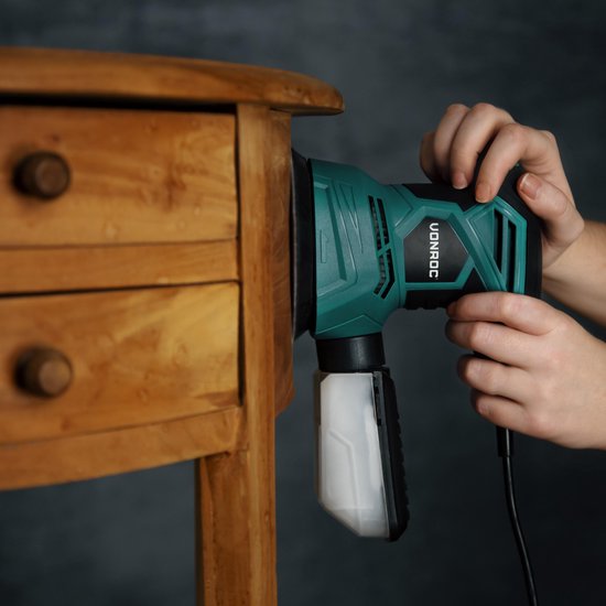 Groene en zwarte excentrische schuurmachine in gebruik op een houten meubel met lades.